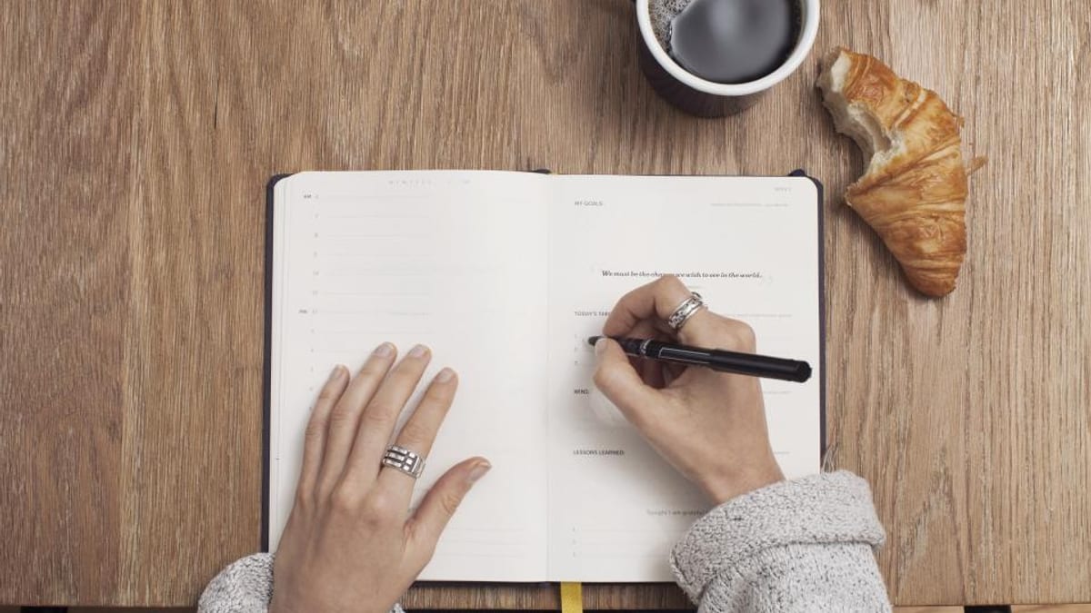 A person writes a plan in an open notebook beside a cup of coffee